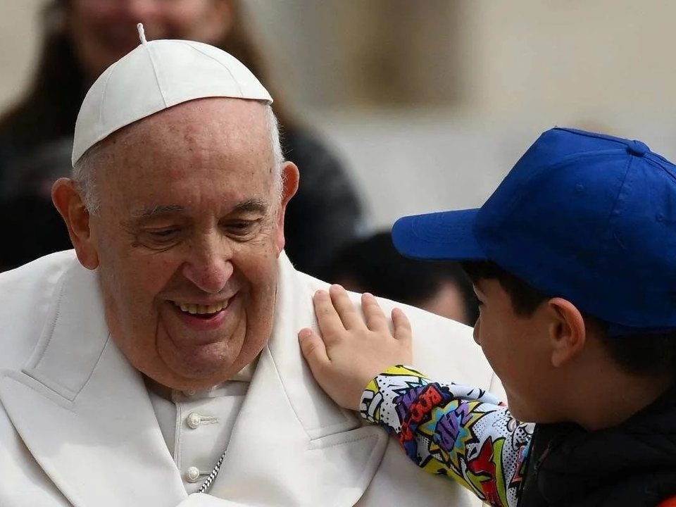 Papa Francisco sorrindo enquanto um menino de boné azul põe a mão no ombro dele